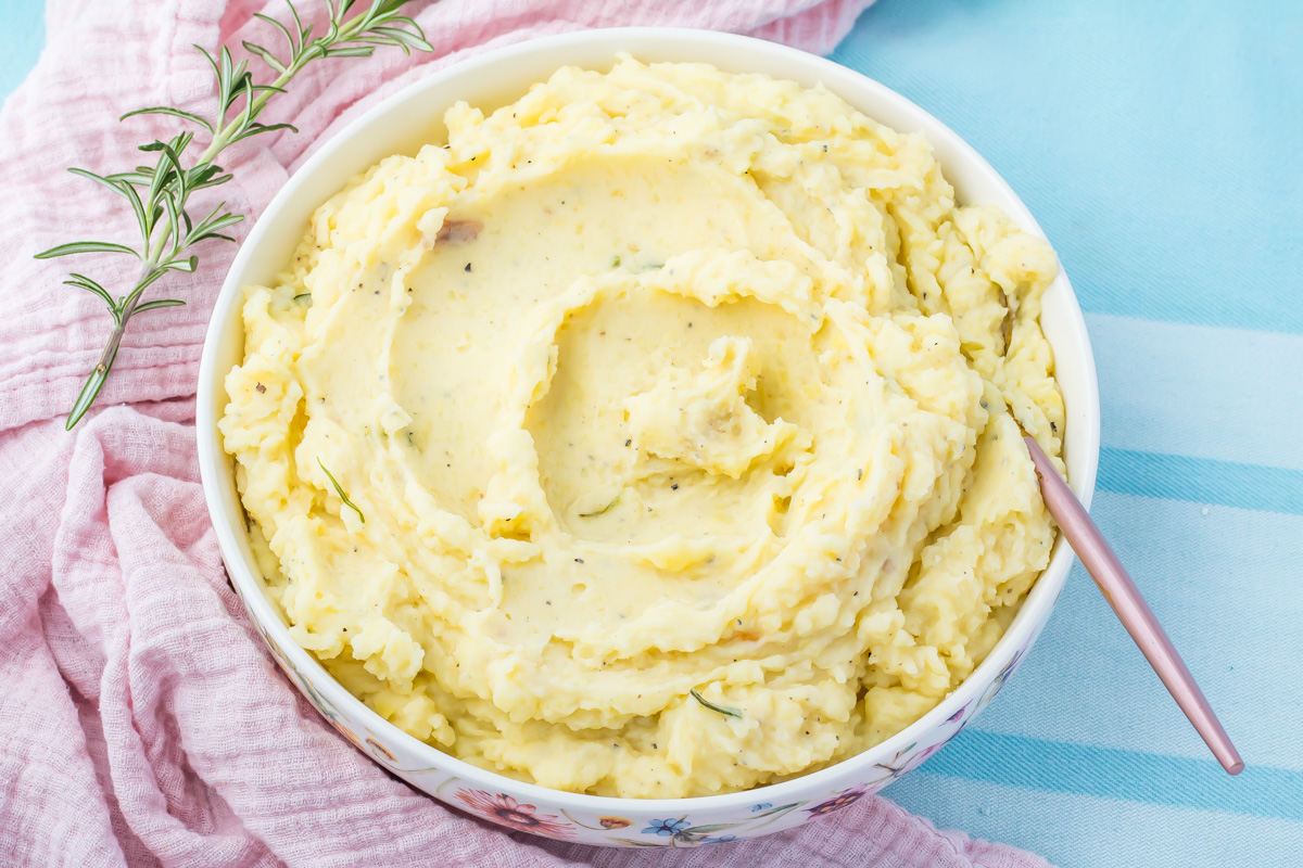 A bowl of cottage cheese mashed potatoes with herbs, placed on a light blue surface next to a pink cloth and a sprig of rosemary, with a spoon in the bowl.