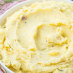 A bowl of creamy cottage cheese mashed potatoes garnished with herbs sits on a pink cloth with a sprig of rosemary nearby.