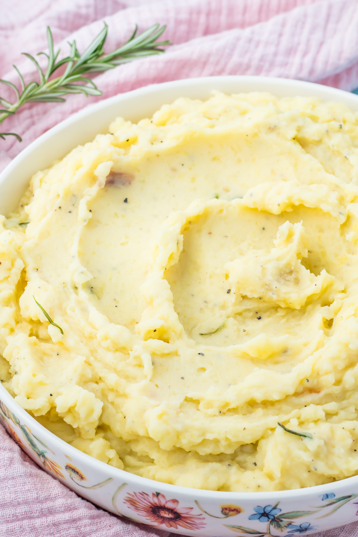 A bowl of creamy cottage cheese mashed potatoes garnished with herbs sits on a pink cloth with a sprig of rosemary nearby.
