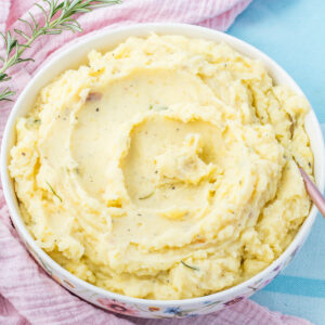 A bowl of creamy cottage cheese mashed potatoes garnished with herbs sits on a pink cloth, accompanied by a sprig of rosemary.