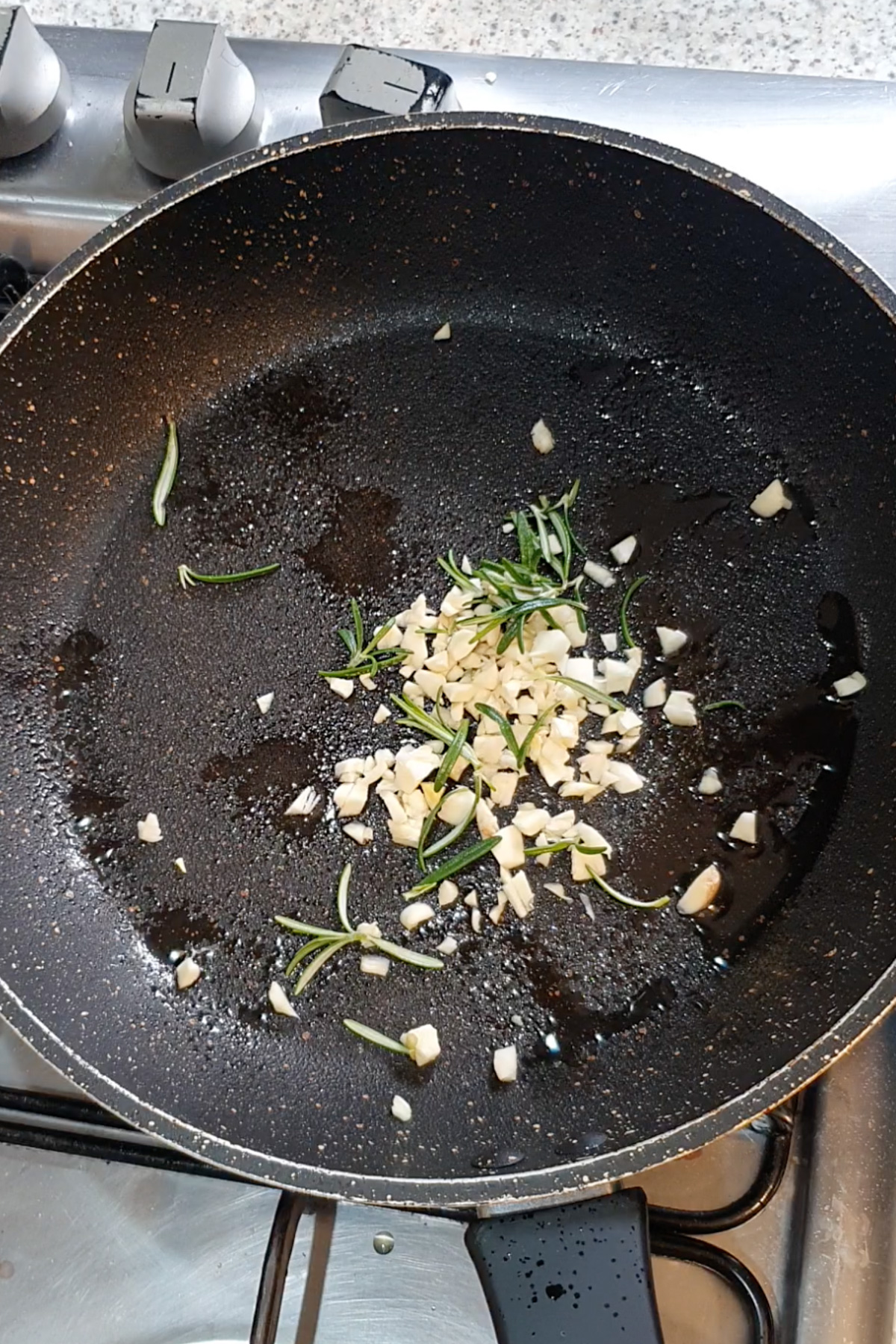 Chopped garlic and rosemary sprigs saut&eacute;ing in olive oil in a nonstick frying pan on a gas stove.