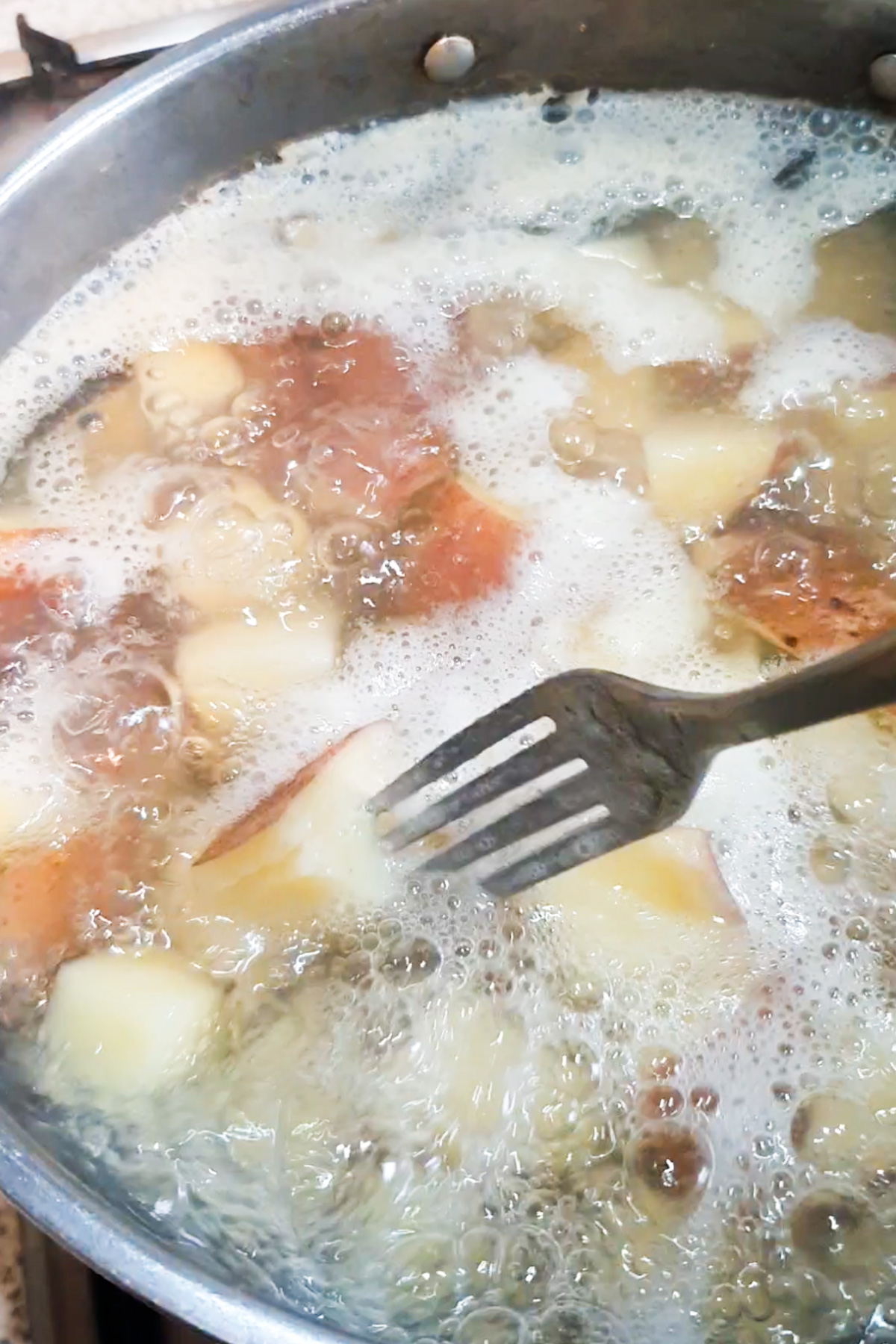 A fork is used to check pieces of potatoes with skin boiling in water in a pot, bubbles and foam on the surface.