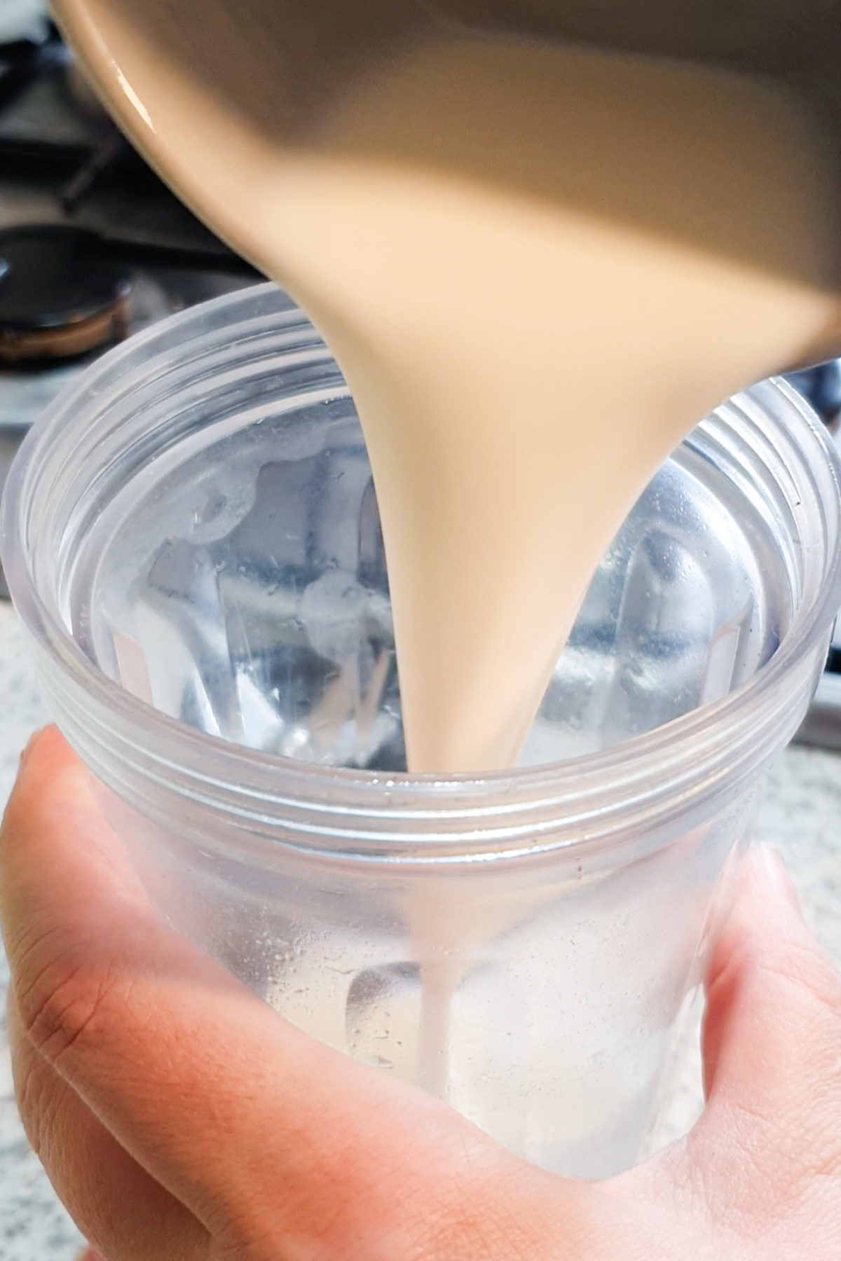 A hand holds a clear plastic cup while a thick, light liquid is being poured into it from a container above.