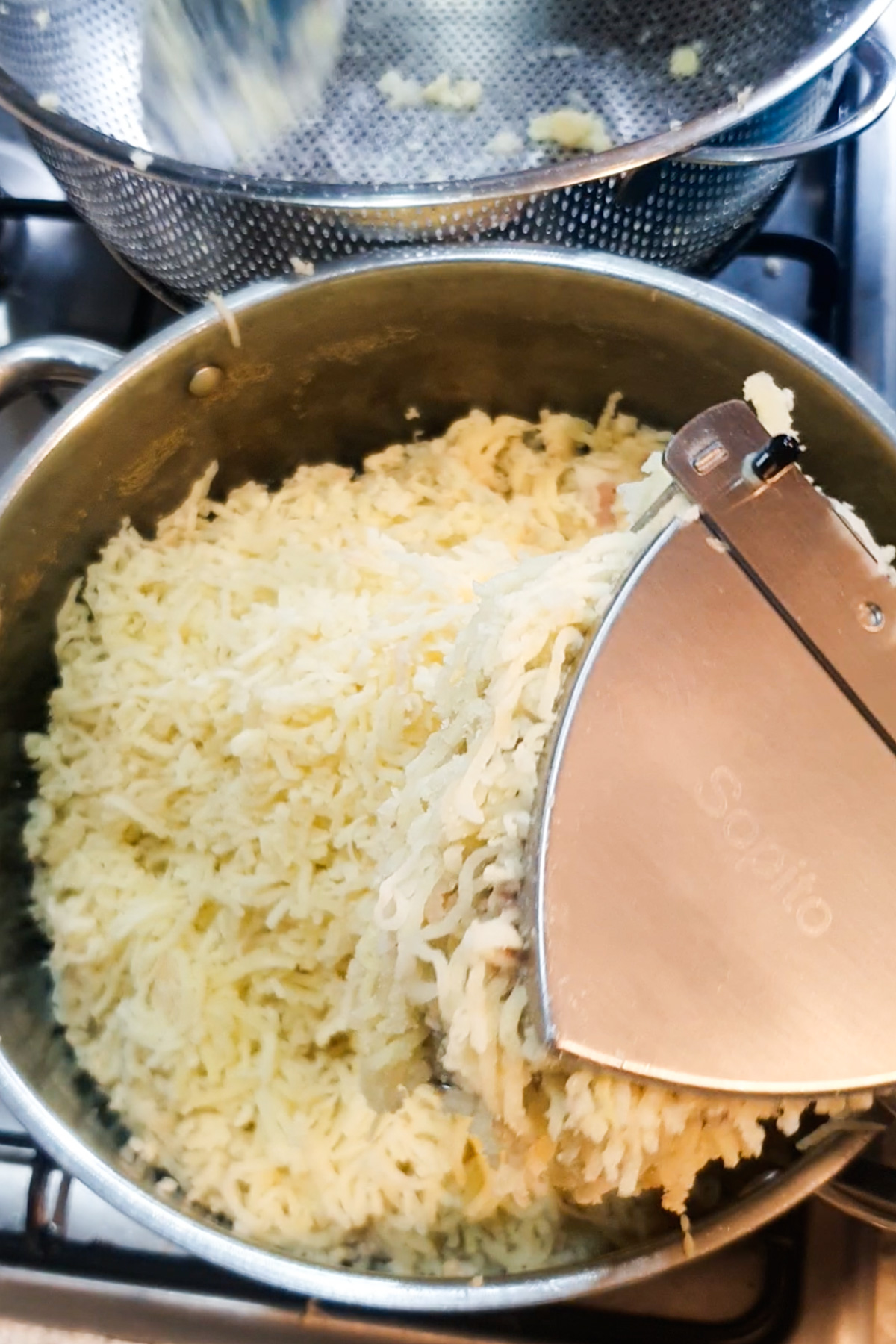 A potato ricer is being used to press cooked potatoes into a pot on a stovetop.