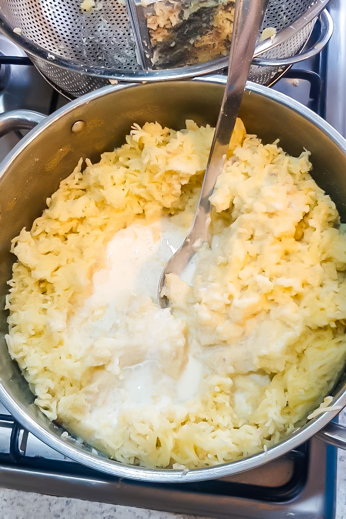 A pot of creamy mashed potatoes with milk being stirred by a metal spoon sits on the stovetop.