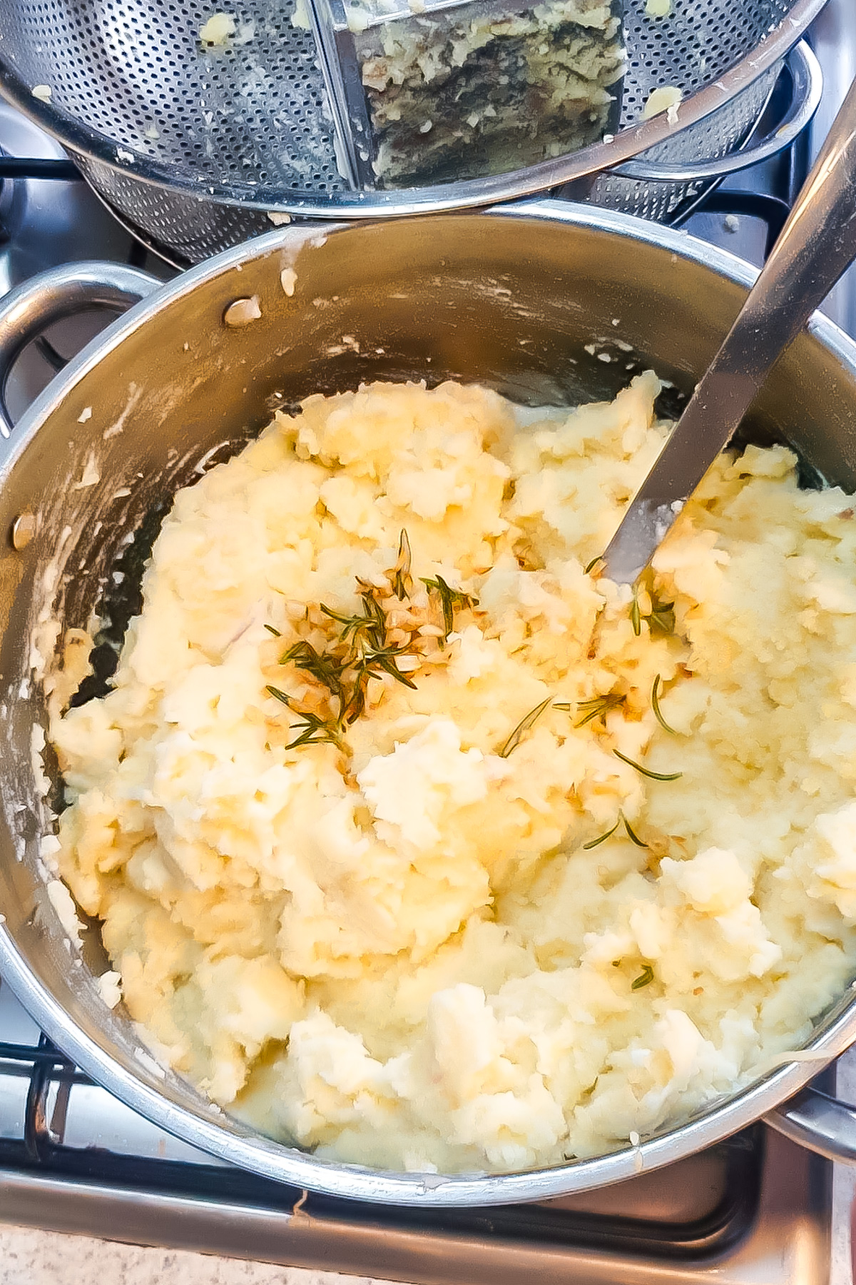 A pot of mashed potatoes with herbs and a metal spoon on a stovetop, with a metal strainer and potato masher in the background.