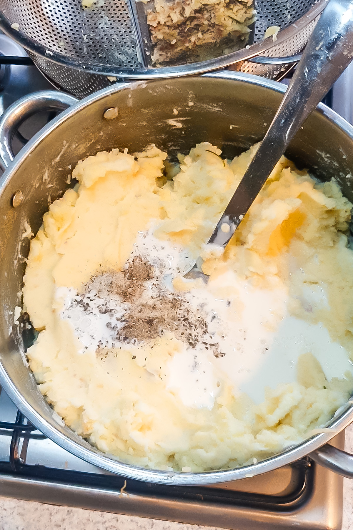 A pot of cottage cheese mashed potatoes on a stove with milk, salt, and pepper being mixed in with a metal spoon.