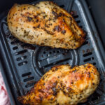 Two seasoned, cooked skinless boneless chicken breasts sit in the basket of an air fryer, making for a perfect air fryer chicken breast meal.