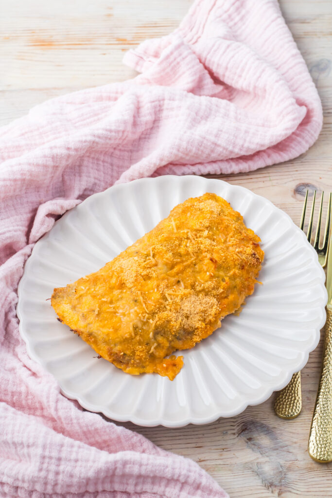 A baked High protein pizza pocket sits on a white scalloped plate next to a gold fork and knife, with a pink cloth napkin on a light wooden surface.