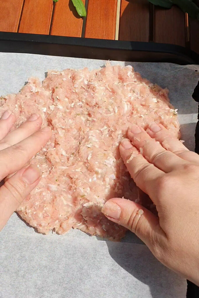 Two hands flatten a high protein raw ground meat mixture on a sheet of parchment paper, preparing it for cooking on a wooden surface—an ideal base for savory pizza pockets.