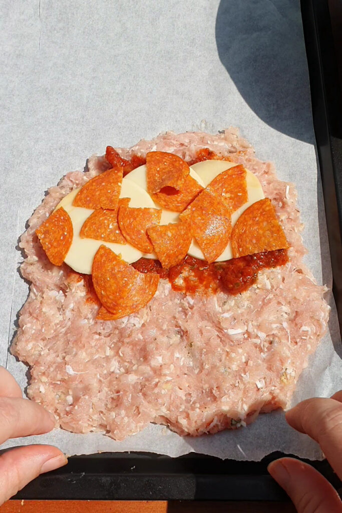 Ground meat is spread on parchment paper, creating a high protein base, with slices of cheese and pepperoni layered on top. Two hands are holding the edges of the parchment, prepping these delicious Pizza Pockets before baking.