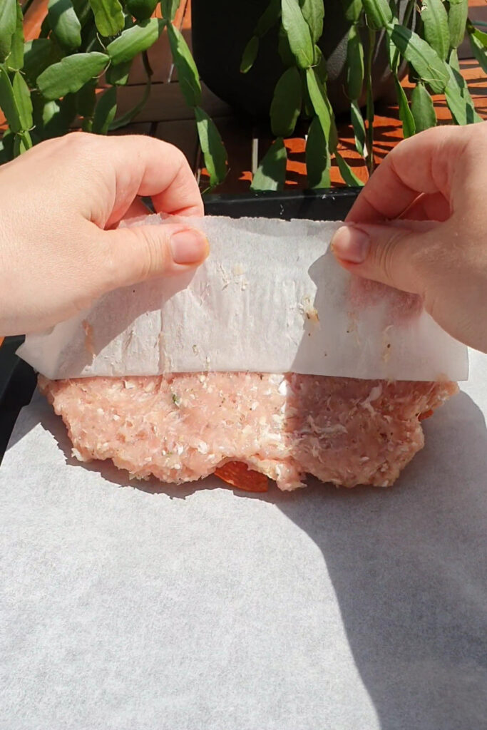 Hands rolling out seasoned high protein ground meat between two sheets of parchment paper, with a plant visible in the background—perfect for making homemade pizza pockets.