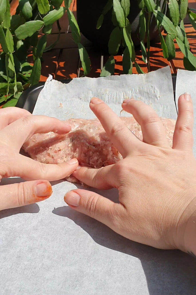 Two hands shape a raw ground meat mixture on a sheet of parchment paper, preparing the perfect base for High Protein Pizza Pockets, with green leafy plants visible in the background.
