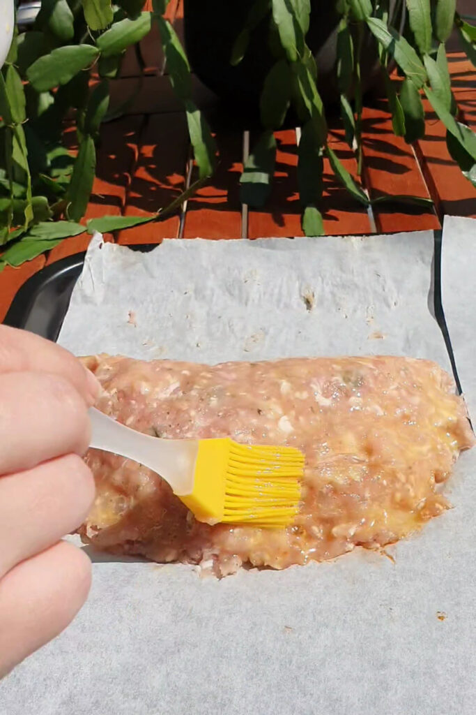A hand uses a yellow silicone brush to spread egg wash over a high protein raw meatloaf mixture on parchment paper outdoors.