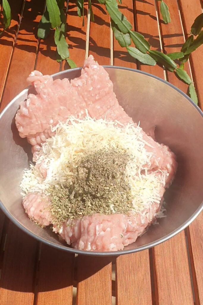 A metal bowl containing raw ground meat topped with shredded cheese and dried herbs sits on a wooden table with leaves in the background—perfect ingredients for crafting a High Protein Pizza or savory Pizza Pockets.