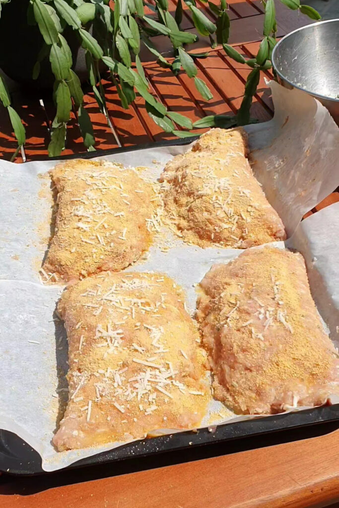 Four breaded chicken thighs on parchment paper atop a baking tray, sprinkled with grated cheese, are set outside on a wooden table next to a green plant—a delicious high protein alternative to classic meals like Protein Pizza or Pizza Pockets.