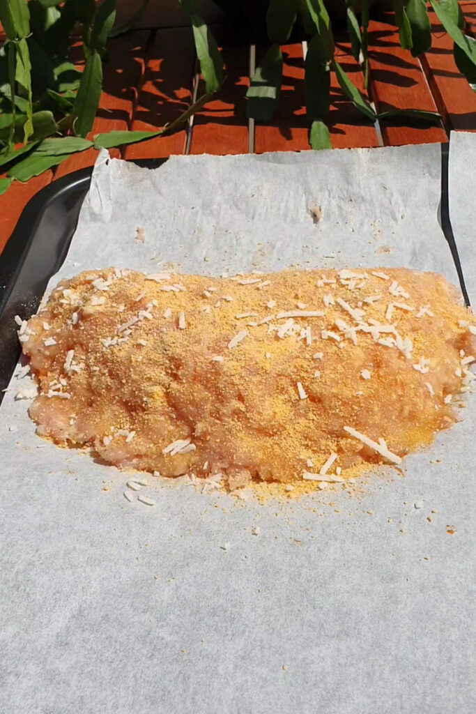 A breaded and cheese-topped raw meatloaf sits on parchment paper on a baking tray, outdoors in sunlight, promising a high protein meal perfect for those who crave hearty flavors.
