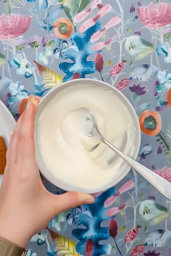 A hand holding a bowl of creamy Greek yogurt protein cheesecake with a spoon, placed on a colorful floral tablecloth.