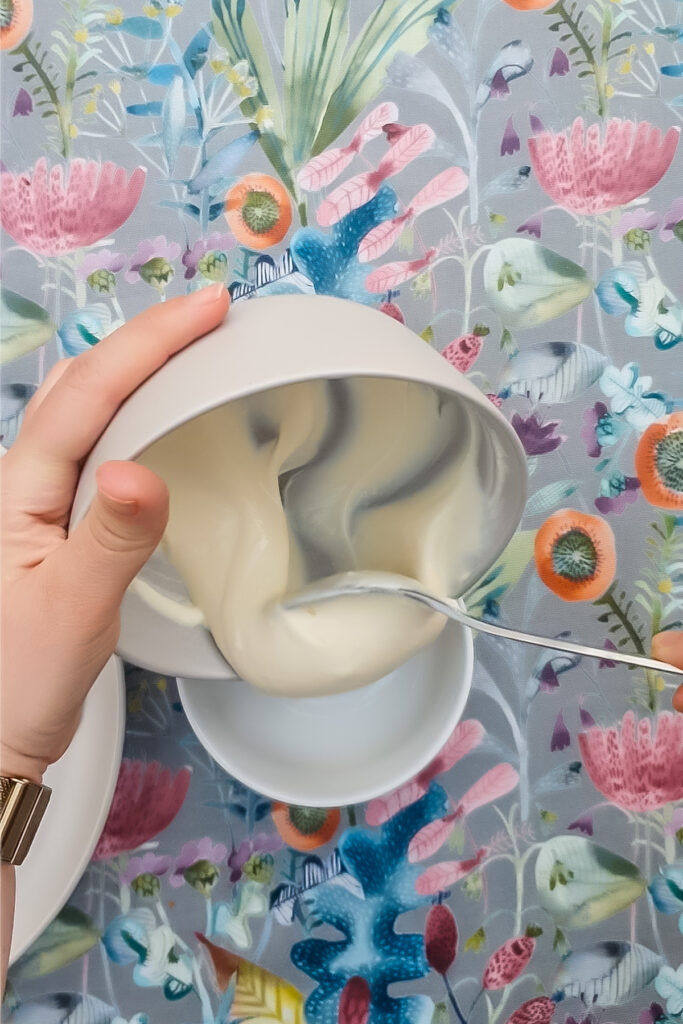 A person uses a spoon to transfer creamy Greek yogurt from one bowl to another, with a floral-patterned tablecloth in the background&mdash;perhaps prepping ingredients for a protein cheesecake.