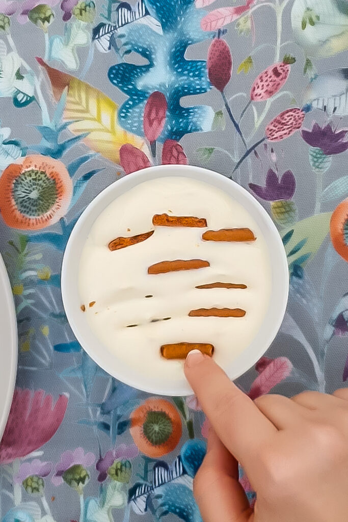 A hand places a stick of cinnamon on a bowl of greek yogurt protein cheesecake, with more cinnamon sticks arranged on top. The bowl sits on a floral-patterned tablecloth.