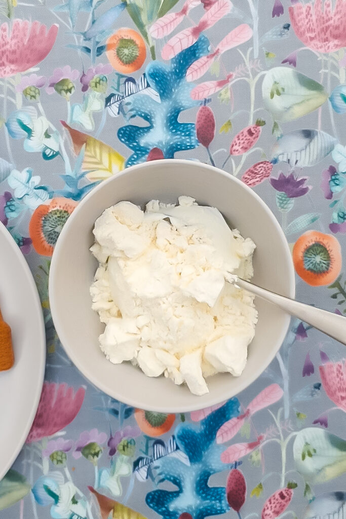 A white bowl filled with scoops of vanilla ice cream and a spoon, placed on a table with a colorful floral-patterned tablecloth&mdash;perfect alongside a slice of greek yogurt protein cheesecake.