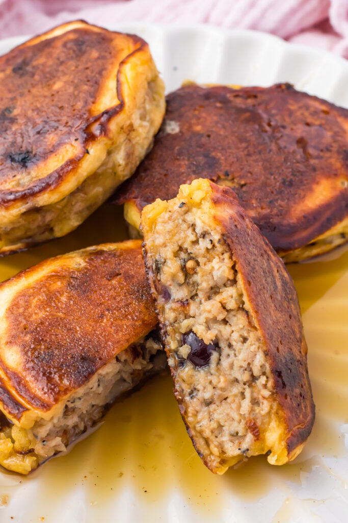 Four browned sausage pancakes, one sliced in half to reveal a moist, textured filling, are arranged on a white plate with visible syrup.