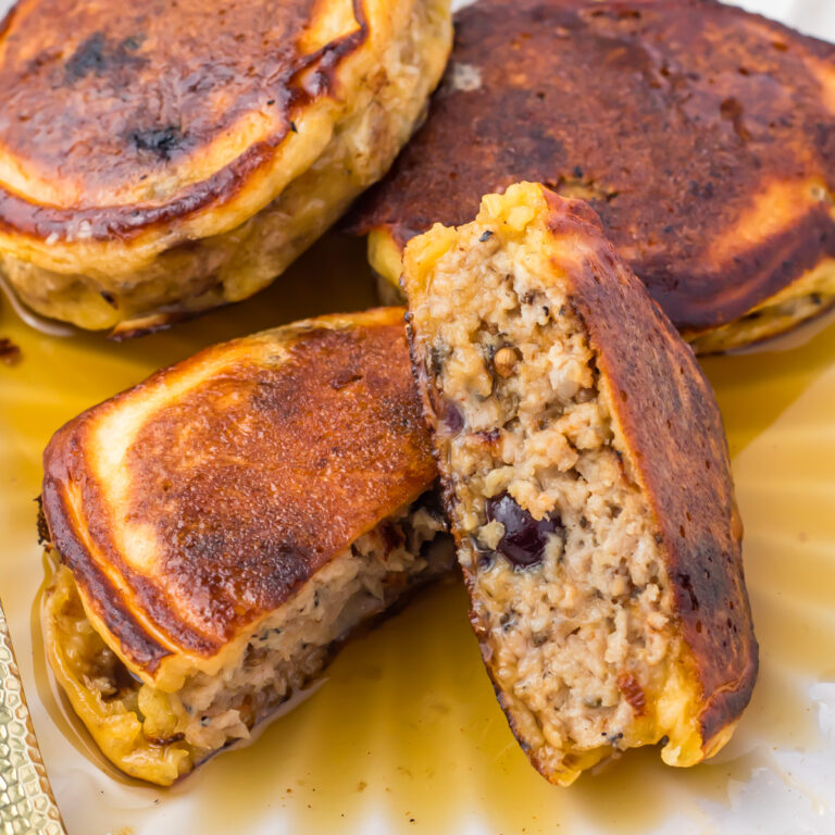Three thick, golden-brown blueberry sausage pancakes are shown on a plate. One pancake is cut in half. Syrup pools underneath.