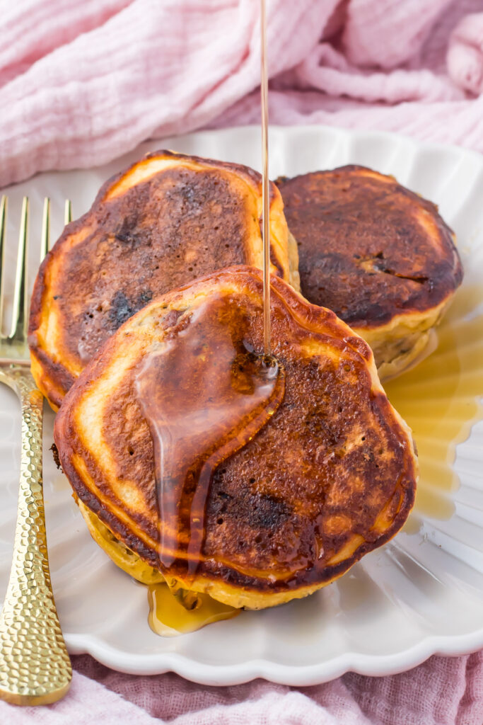 Three blueberry pancakes on a white plate with syrup being poured on top, next to a gold-colored fork and a pink cloth in the background.