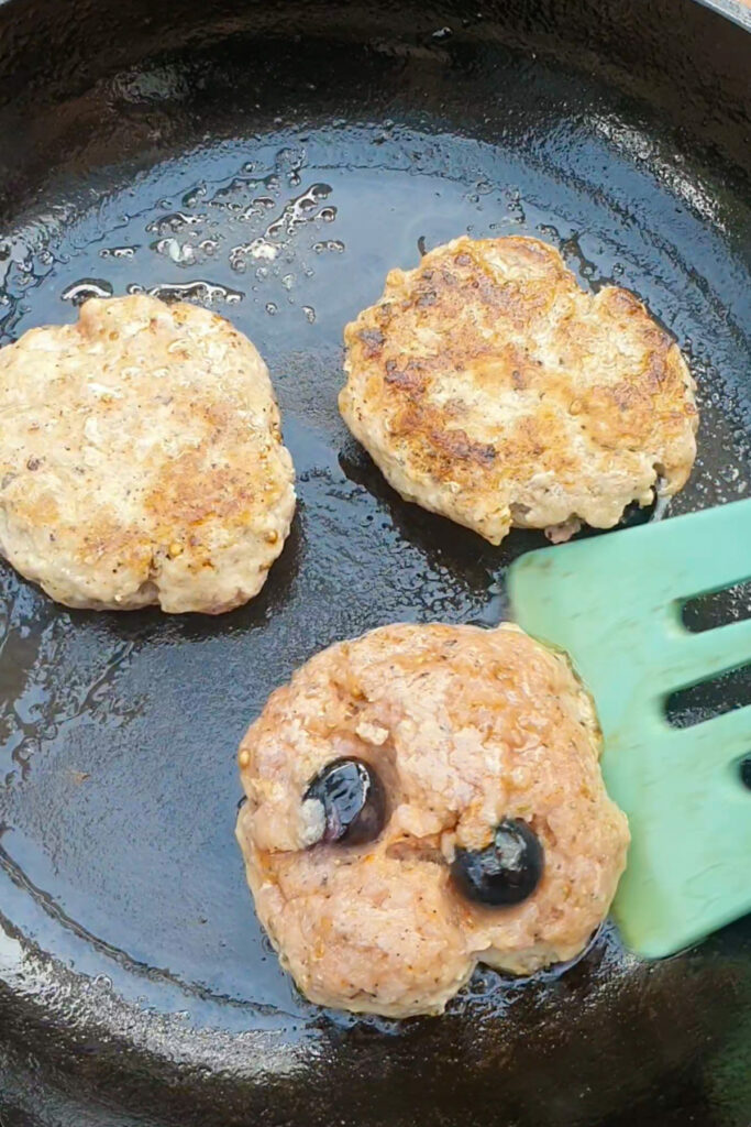 Three sausage patties cooking in a skillet, one being flipped with a green spatula. The patty being flipped contains two blueberries.