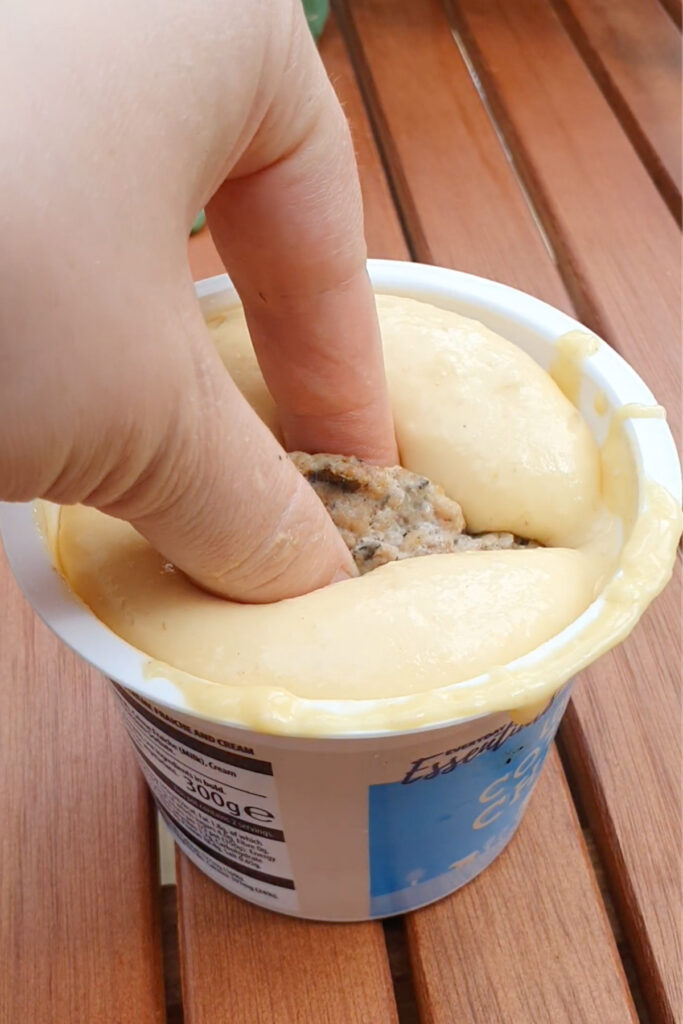 A hand dips two fingers holding a sausage into a cup of pancake batter, with some batter around the rim, on a wooden surface.