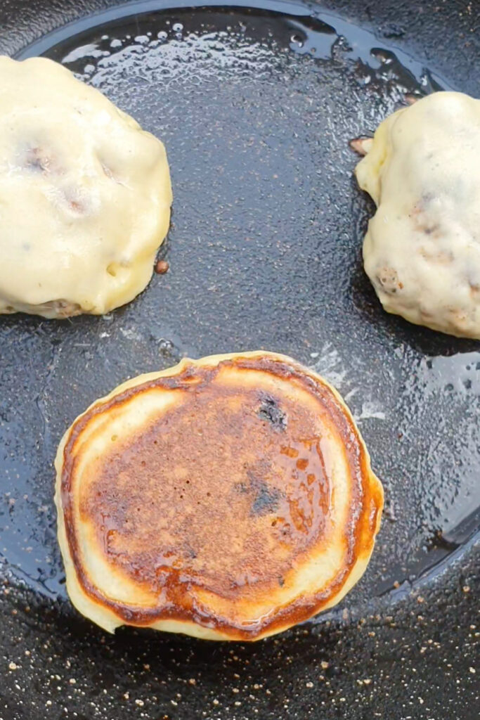 Three sausage pancakes cooking in a frying pan; two haven't been flipped yet and one is flipped showing a browned crust.