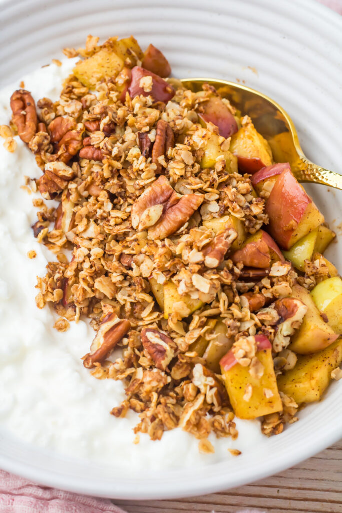 An apple crumble cottage cheese bowl topped with diced apples, granola, pecans, and served with a gold spoon on a white plate.