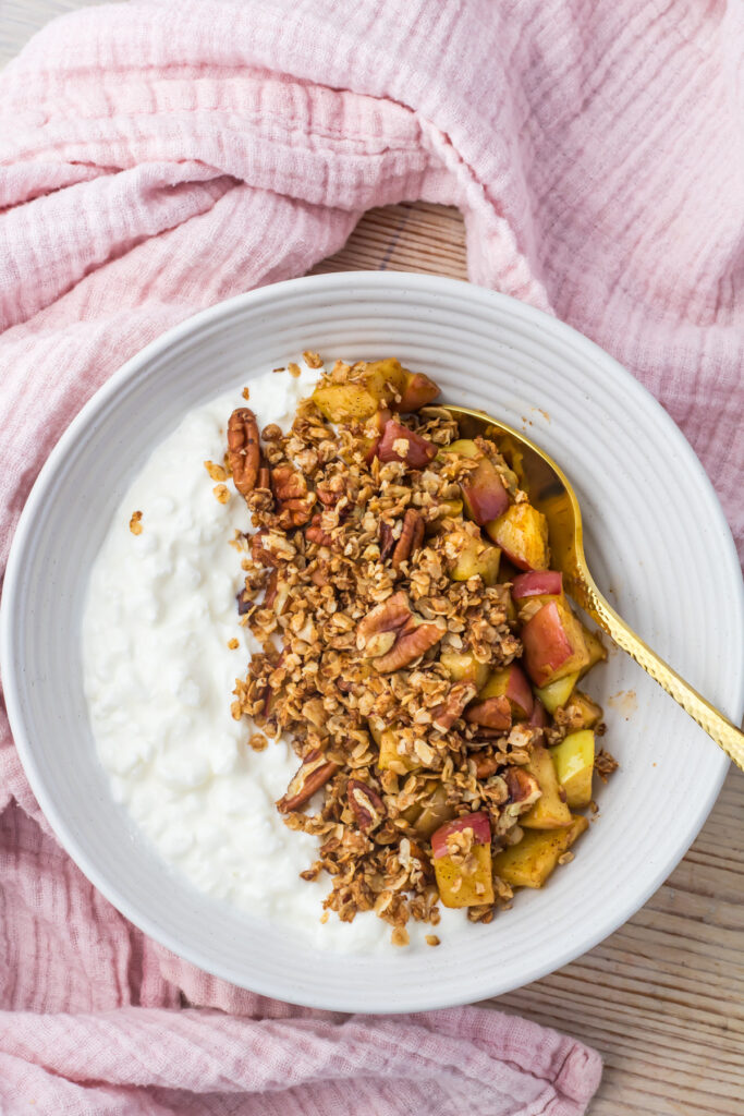 Apple crumble cottage cheese bowl in a white bowl with creamy cottage cheese and apple crumble sits on a wooden table with a pink cloth, with a gold spoon resting inside.