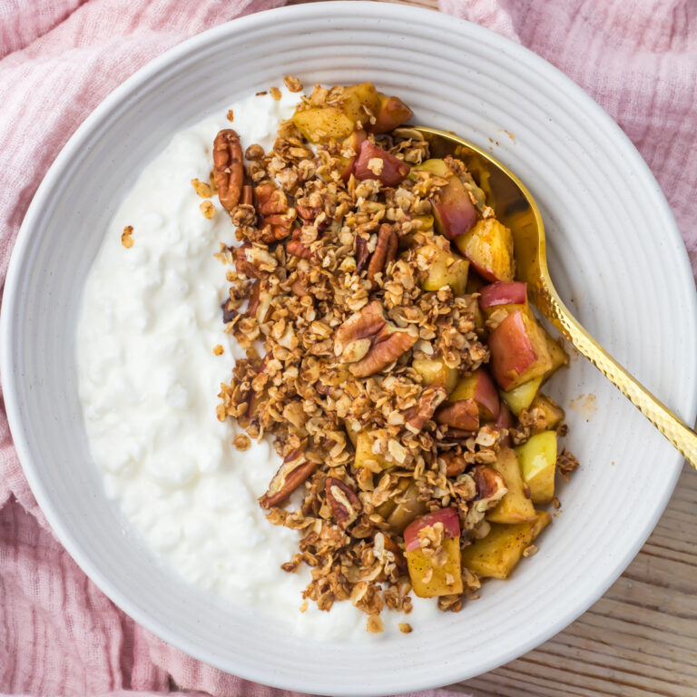 Apple crumble cottage cheese bowl in a white bowl with creamy cottage cheese and apple crumble sits on a wooden table with a pink cloth, with a gold spoon resting inside.