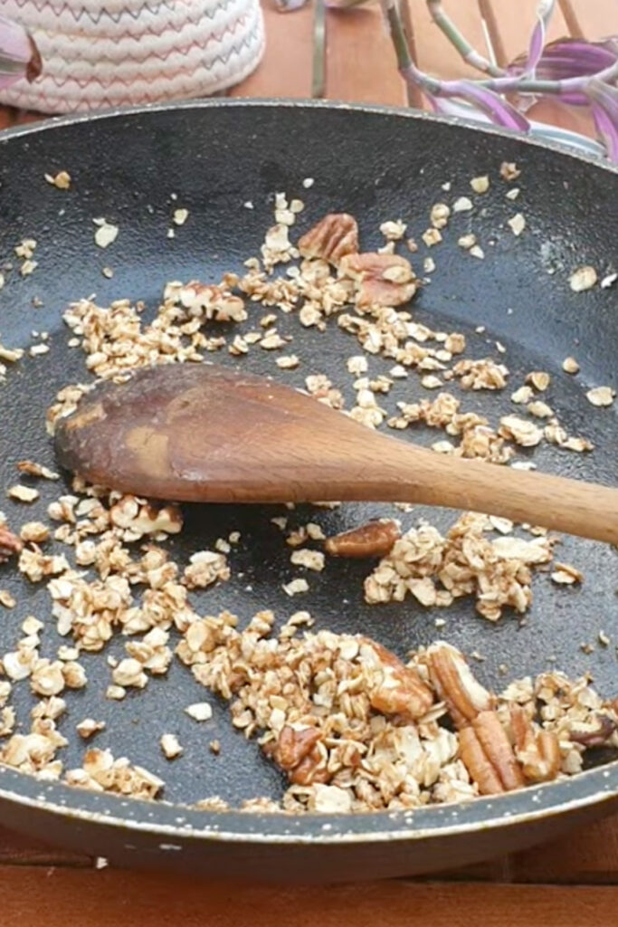 A wooden spoon rests in a frying pan containing toasted oats and pecans on a wooden table.