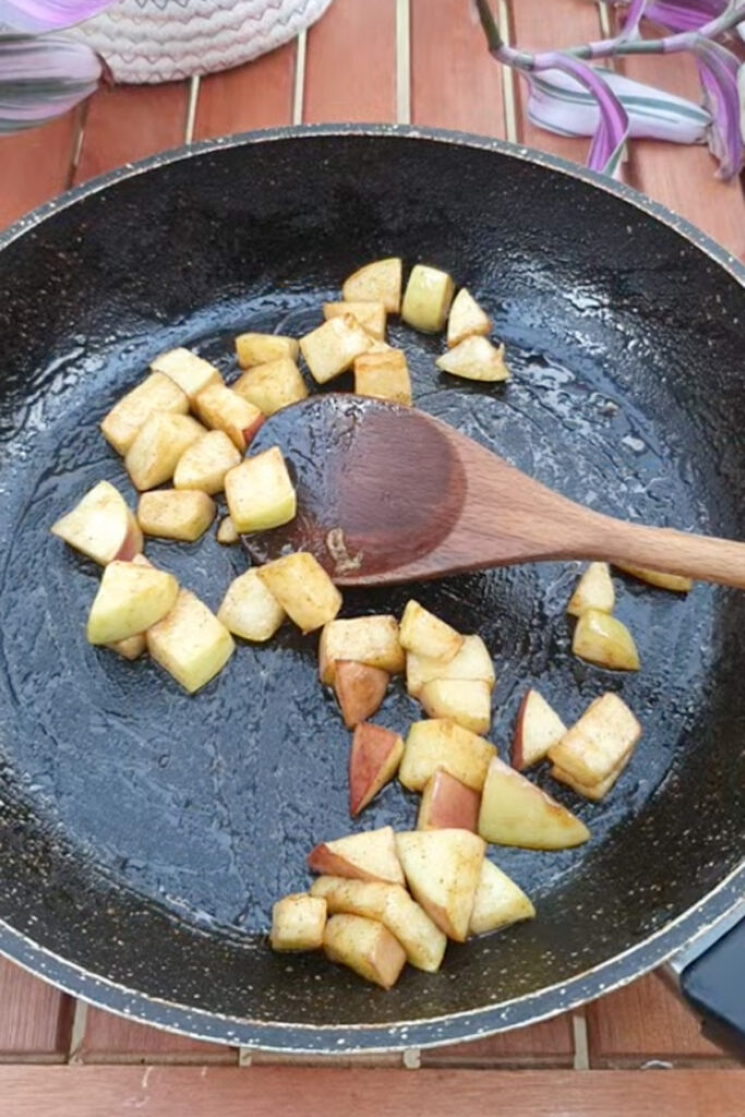 A frying pan with chopped apples being saut&eacute;ed, stirred by a wooden spoon on a wooden surface.