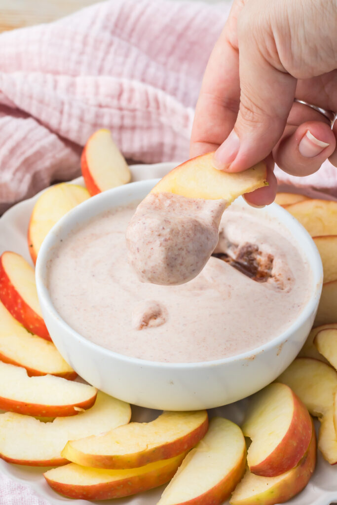 A hand dips a sliced apple into a bowl of creamy, cottage cheese apple dip, with more apple slices arranged around the bowl on a plate.