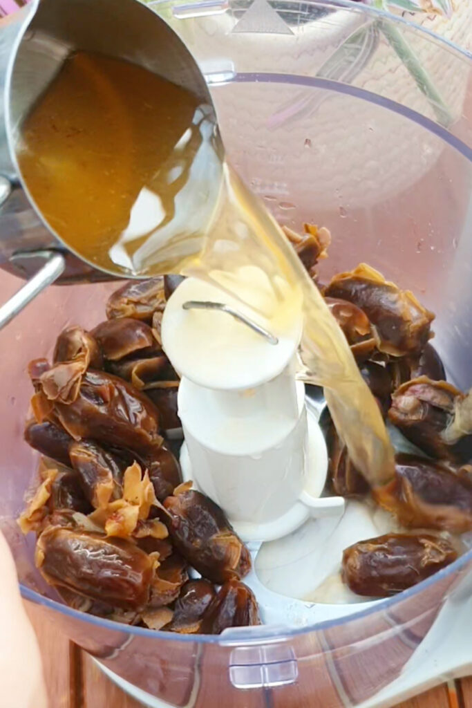 A liquid is being poured from a metal container into a food processor filled with pitted dates.