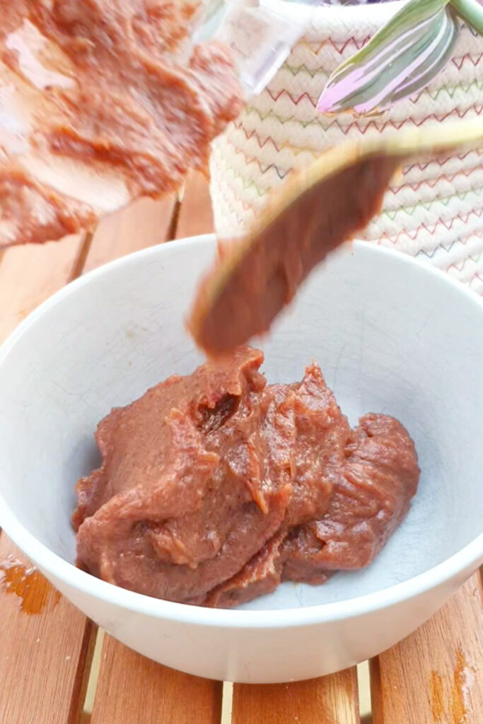 Thick date paste is being scooped into a white bowl on a wooden surface, with a textured cloth and plant in the background.