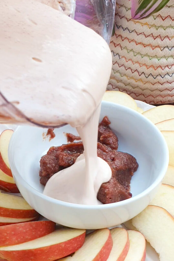 A creamy cottage cheese mixture is being poured into a bowl containing a brown date paste, surrounded by sliced apples on a plate.
