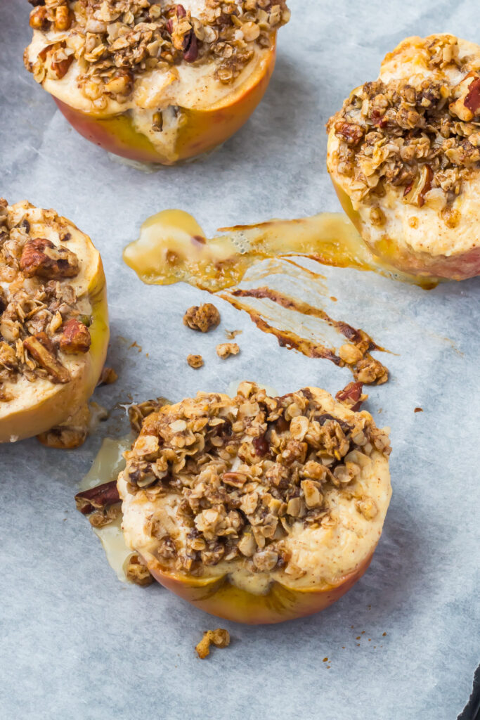 Four cottage cheese baked apples topped with oat and nut crumble are arranged on parchment paper, with melted filling and crumbs visible around them.
