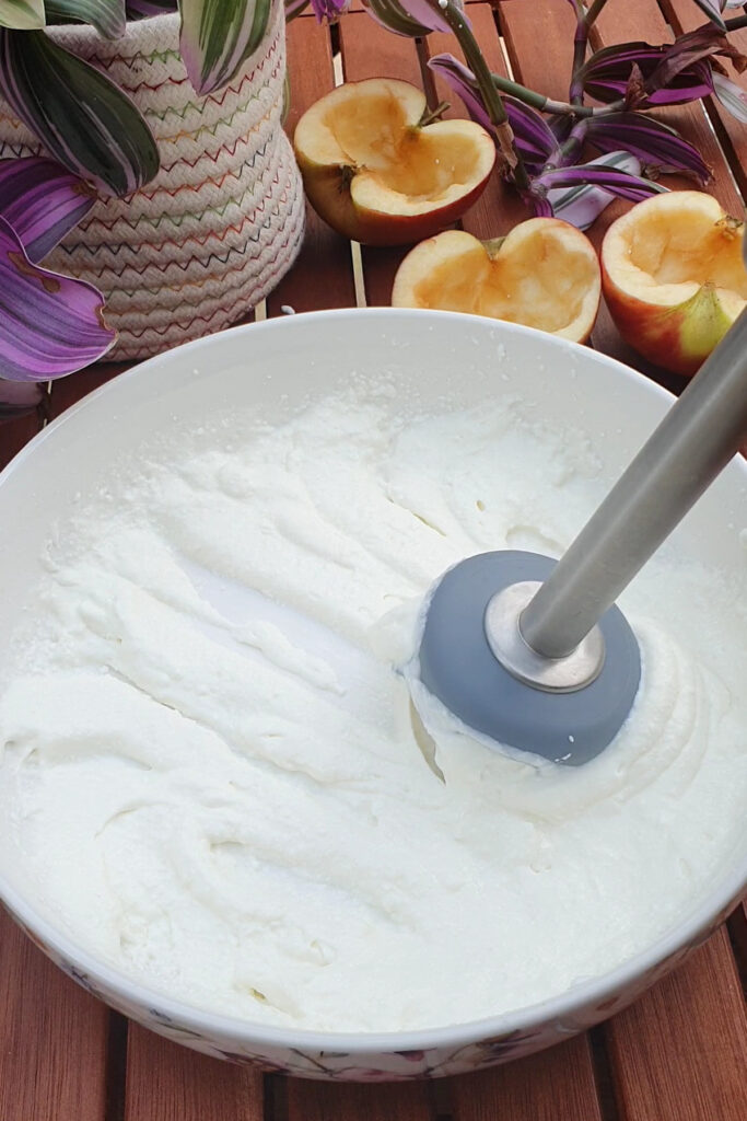 A stick blender is mixing cottage cheese in a white bowl on a wooden table, with halved apples and a potted plant in the background.