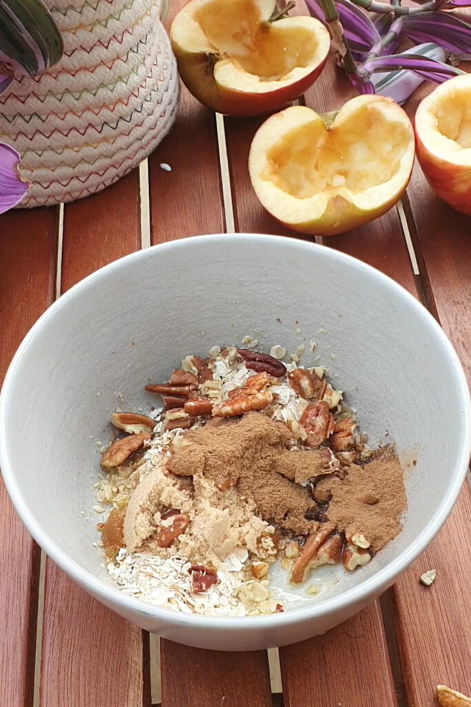 A white bowl with oats, brown sugar, chopped pecans, and cinnamon sits on a wooden table next to cored apple halves and a potted plant.