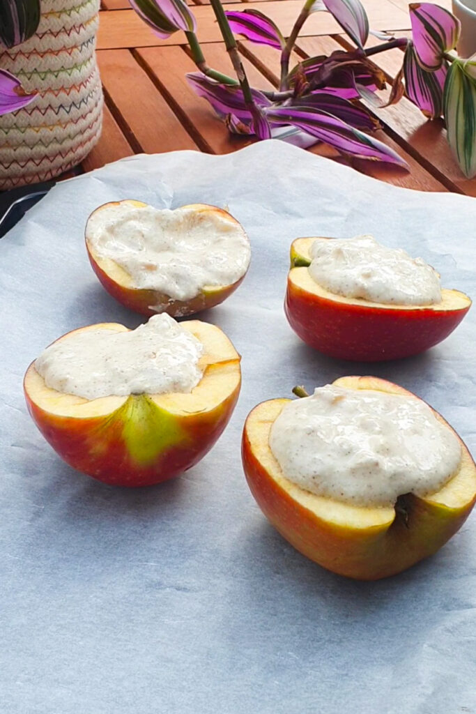 Four baked apple halves filled with a creamy cottage cheese mixture are placed on parchment paper, with a potted plant and a wooden surface visible in the background.