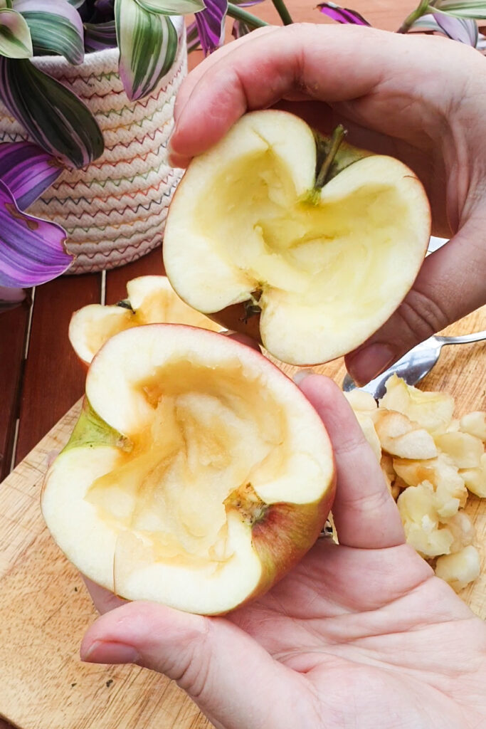 A person holds two halves of a hollowed-out apple above a cutting board, with apple pieces and a potted plant visible in the background.