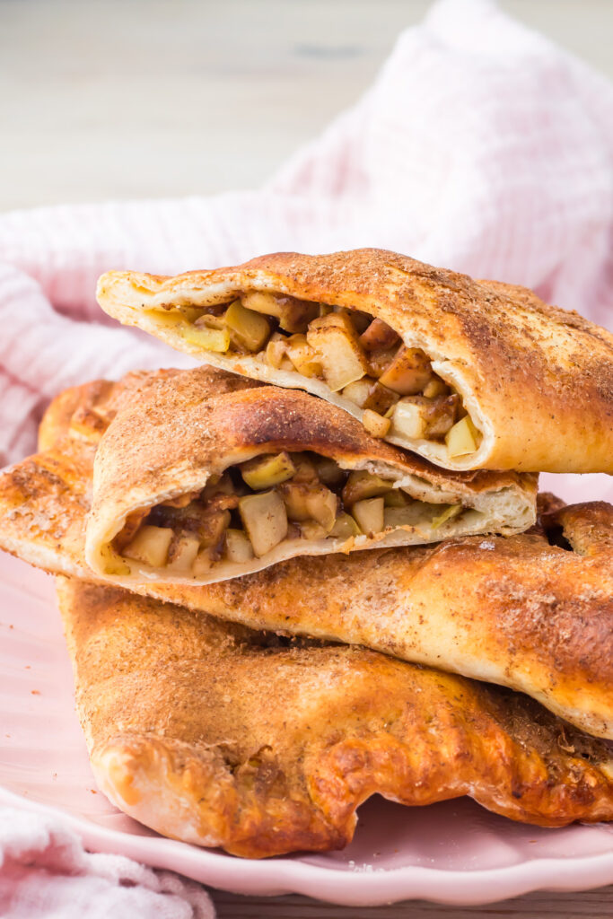 Two golden-brown high protein copycat McDonald&rsquo;s Apple Pies, are filled with chopped apples and cinnamon, stacked on a pink plate with a pink cloth in the background.