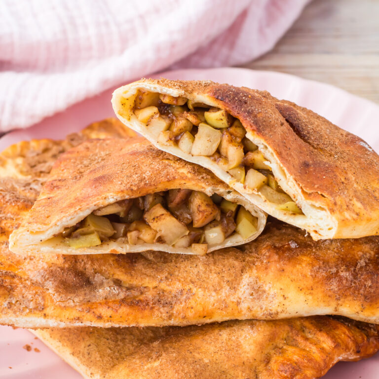 A copycat high protein mcDonad's Apple Pie cut in half, revealing a filling of diced apples and spices, is stacked on a pink plate, with a light cloth in the background.