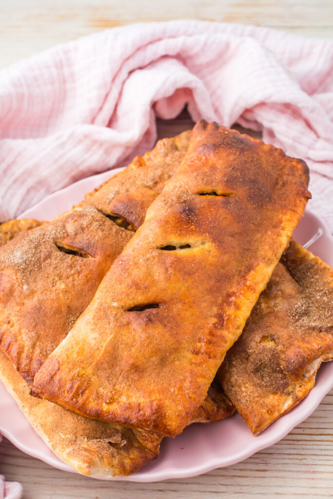 Three golden-brown rectangular high protein copycat McDonald&rsquo;s apple pies, are stacked on a pink plate with a light pink cloth in the background.