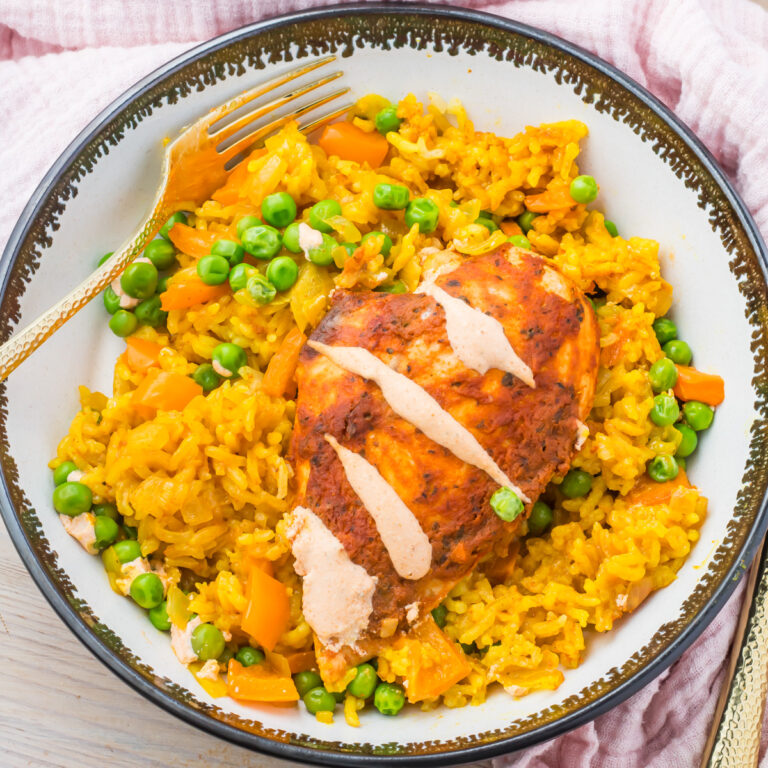 Peri peri chicken and rice with a creamy drizzle served over yellow rice mixed with green peas and diced vegetables in a patterned bowl.