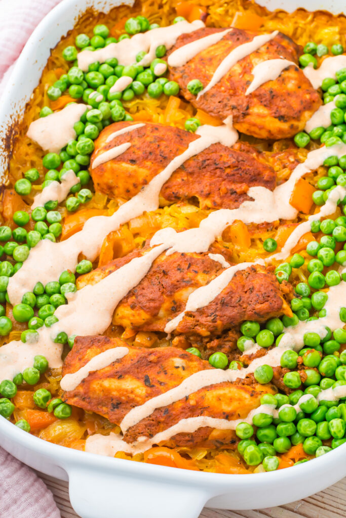 A white baking dish filled with peri peri chicken and rice, featuring spiced chicken breasts, green peas, and diced vegetables drizzled with a creamy sauce.
